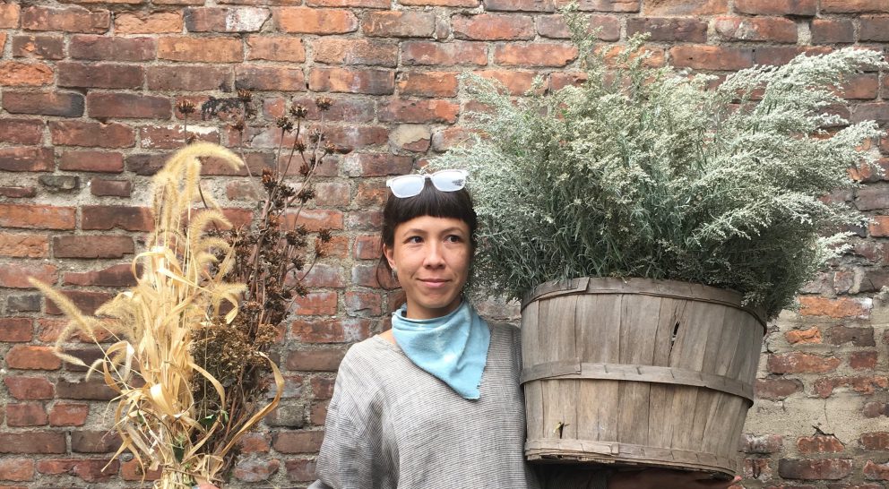 A landscape photo of Azuré Kauikeolani Iversen-Keahi, a light-skinned person holding a large potted plant wearing a grey sweater, light blue scarf and glasses on top of their head. They have dark brown hair with bangs falling in front of their face and the rest pulled back. The background is a brick wall.
