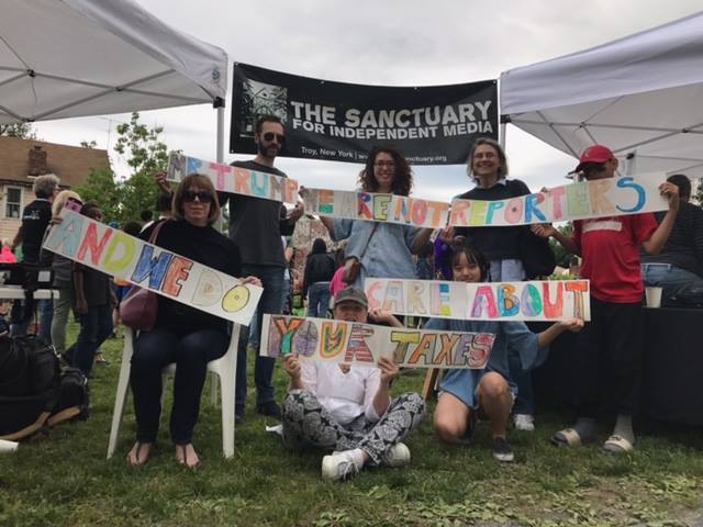 A group of people sitting and standing in front of a Sanctuary for Independent Media sign, holding various letter signs spelling out a message.