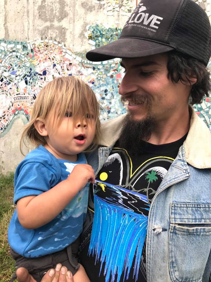 A man is holding a child in his arm, the child is smiling with long blonde hair and he is smiling down at the child.