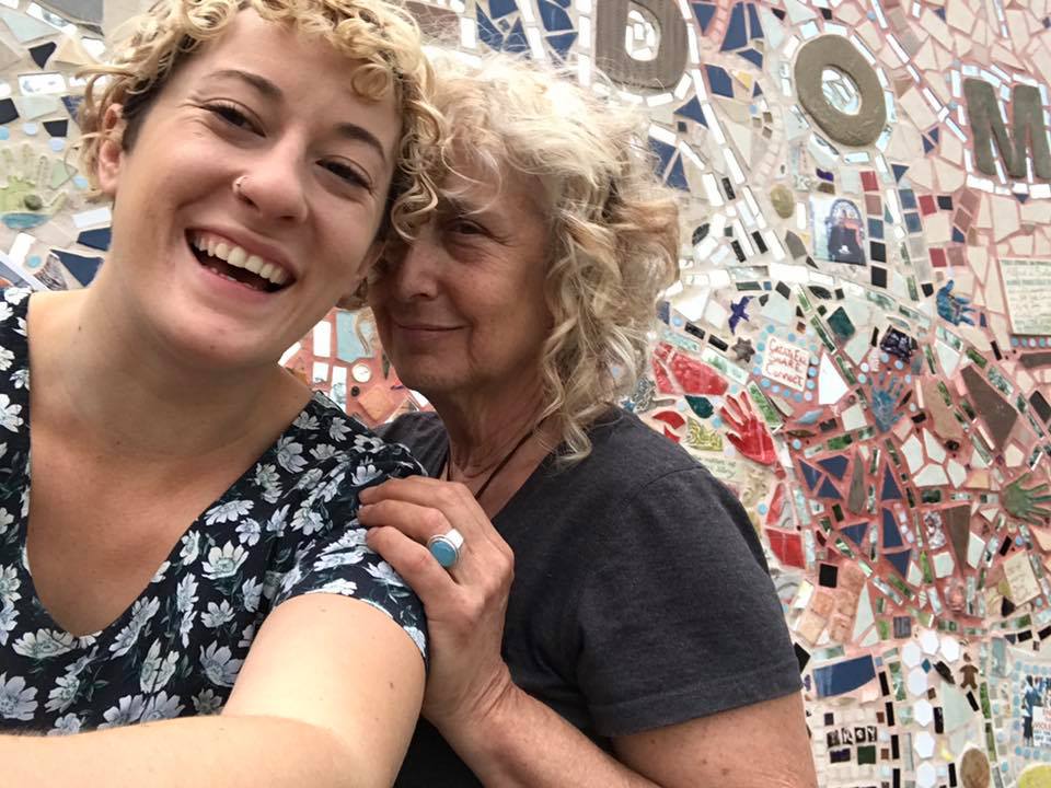 Two peoples standing in front of a mosaic wall, both with curly blonde hair. The one on the left is smiling wide while the other has a less intense smile- both look happy.