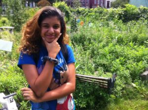 A brown-skinned teenage girl, standing in the Collard City Growers garden, smiles proudly with her hand in a thoughtful position on her chin. 