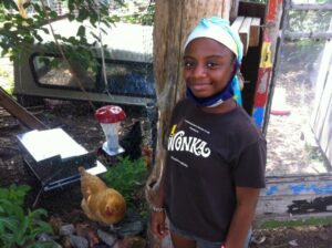 A Black teenage girl, wearing a T-shirt and head wrap, smiles at the camera in the Sanctuary's community garden. 