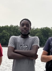 A Black teenage boy in a grey T-shirt stands at the edge of a river with his arms crossed. 