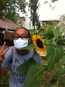 A Black teenage girl with glasses poses next to a sunflower with her hand making a peace sign. 