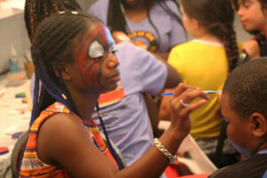 Genesis Cooper, whose face is painted like Spider-Man's mask, applies face paint to a dark-skinned boy. 