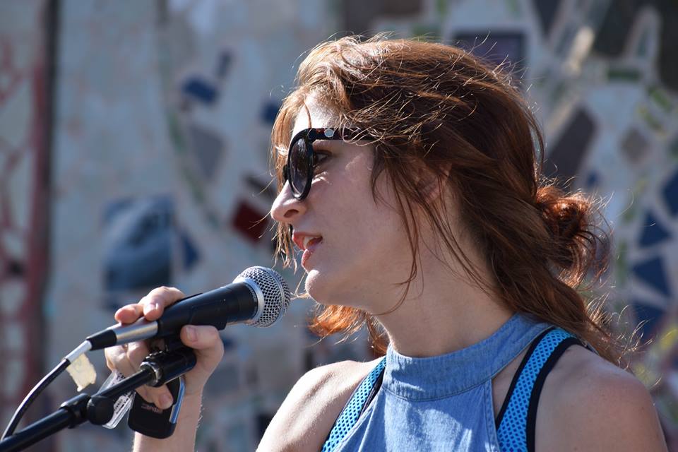 A light-skinned woman with long red hair is giving a speech into the microphone, looking into the distance.