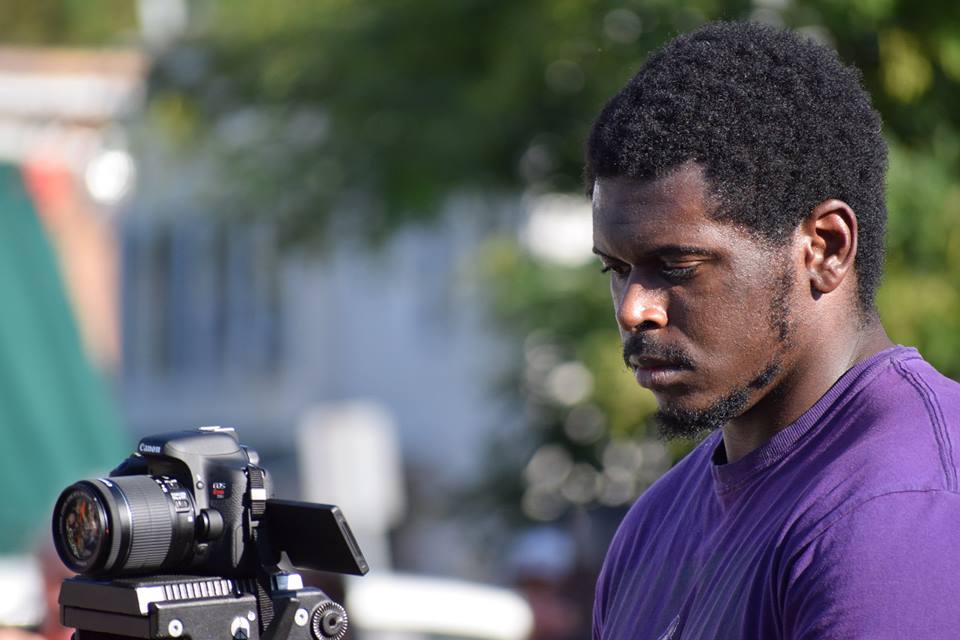 A man is standing behind a camera, focusing on the events of the festival. He is wearing a purple shirt and has dark skin with black hair.