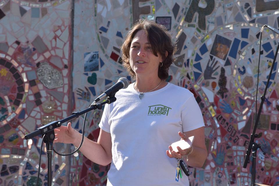 A woman standing in front of a mosaic wall wearing a white shirt, giving a speech into a microphone.