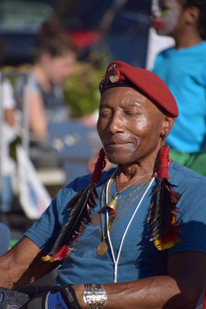 A person of color sitting with their eyes closed, wearing a red hat with feather accessories.