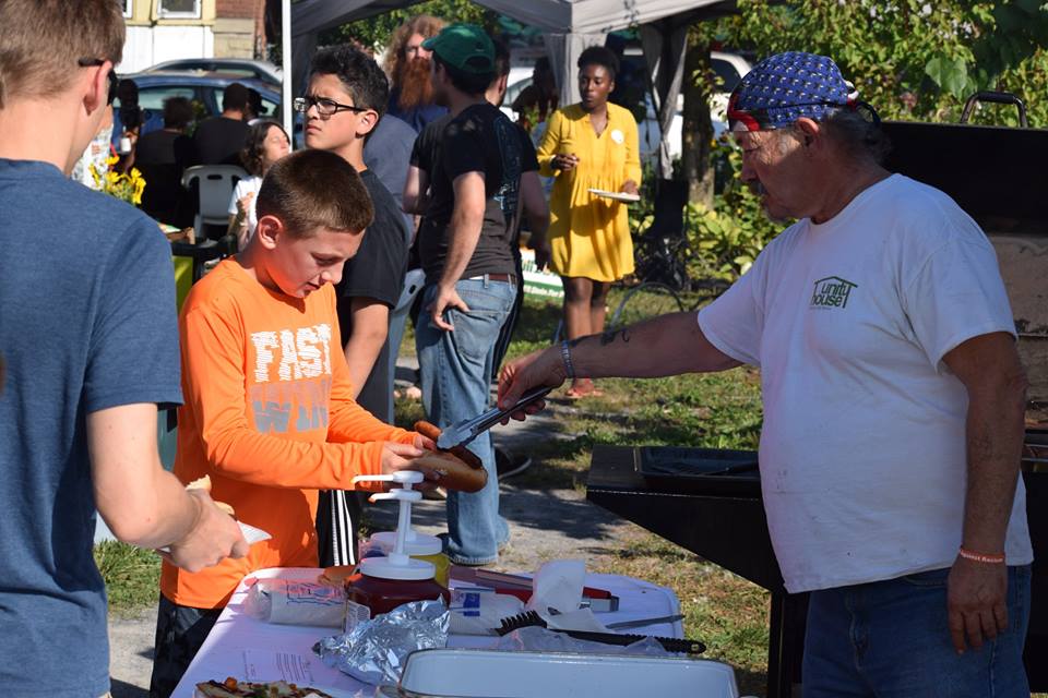An older man is serving a young teenager a hot dog at the festival, with others gathered in the background.