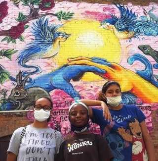 Shansanique Pollack, Genesis Cooper, and Gabby Espada pose in front of a colorful mural. The mural depicts two hands, one blue and one orange, clasping in front of the sun, surrounded by plants and animals.