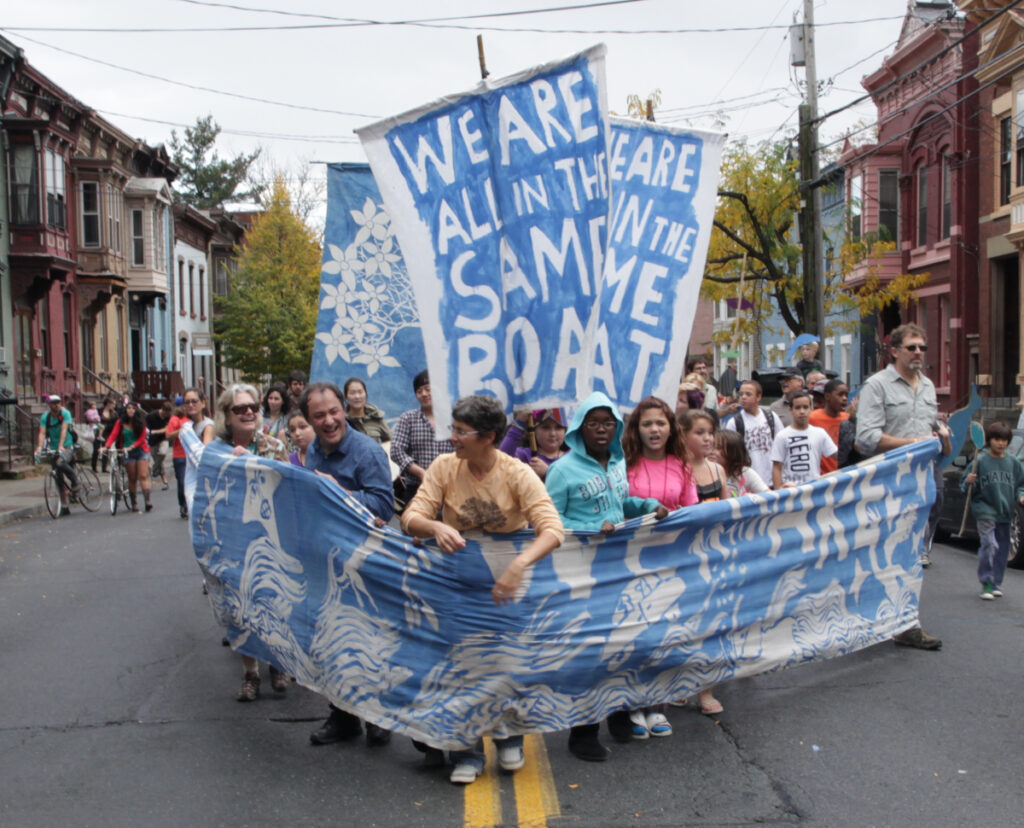 Bread and Puppet march down 6th Ave with banners and flags saying "we're all in the same boat"