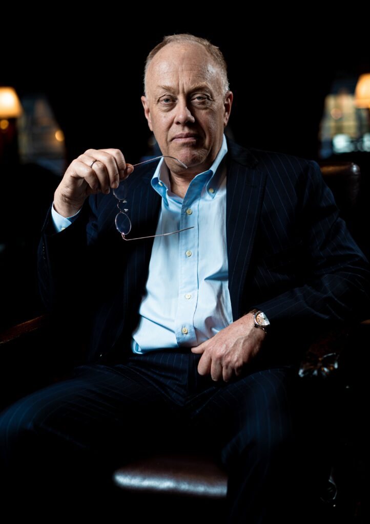 Medium-framed shot of Chris Hedges wearing a light blue button up and black blazer, holding his glasses.