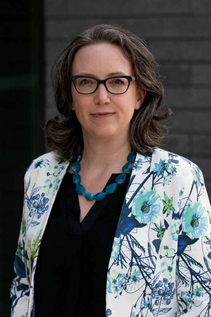 A head shot of Dr. Jennifer Willet, a light-skinned woman with medium-length brown hair wearing glasses and a floral blazer over a black blouse.