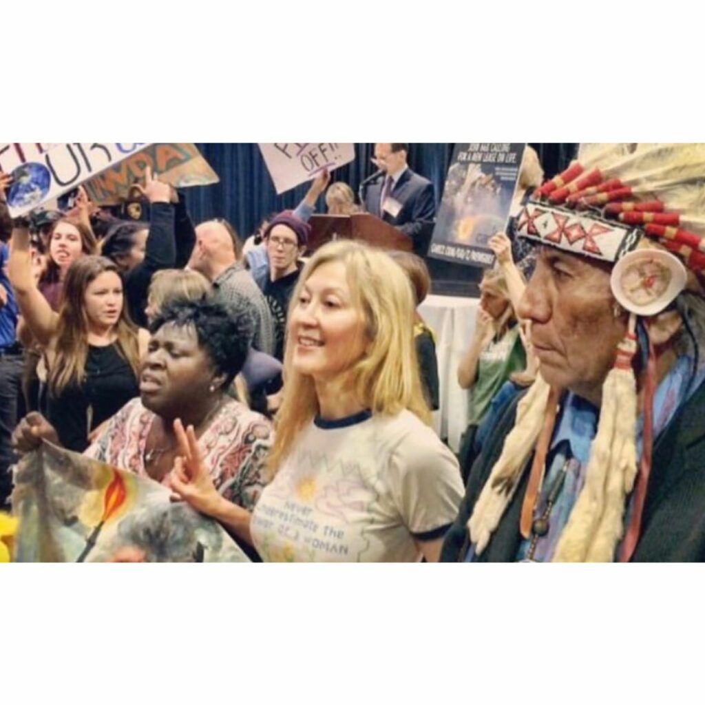 Janet MacGillivray is in a crowd between an Indigenous American man on her left with a large feather headdress and an Aboriginal woman on her right. Janet MacGillivray has long, blond hair and is giving the peace sign.