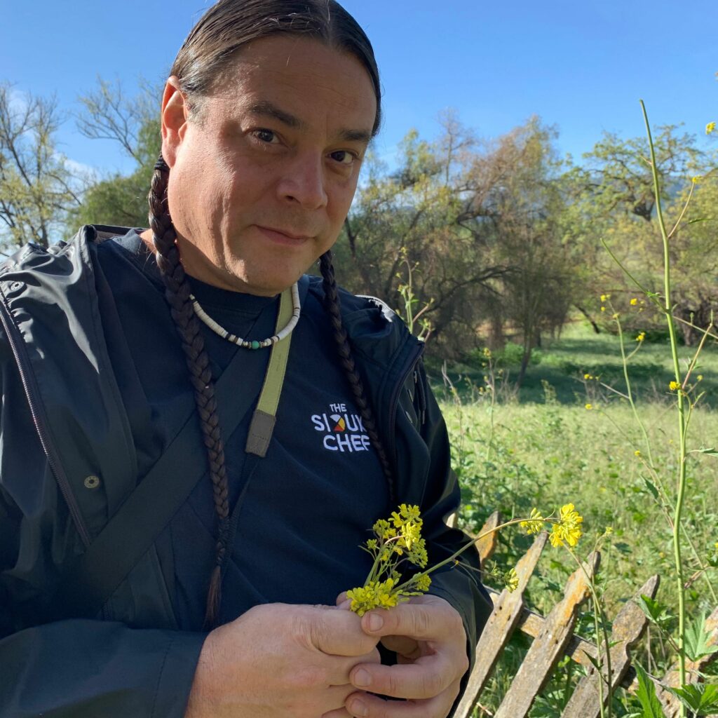Chef Sean Sherman is outdoors with yellow flowers in his hand. He is wearing a Sioux Chef tshirt, wears two braids, and a thin beaded necklace