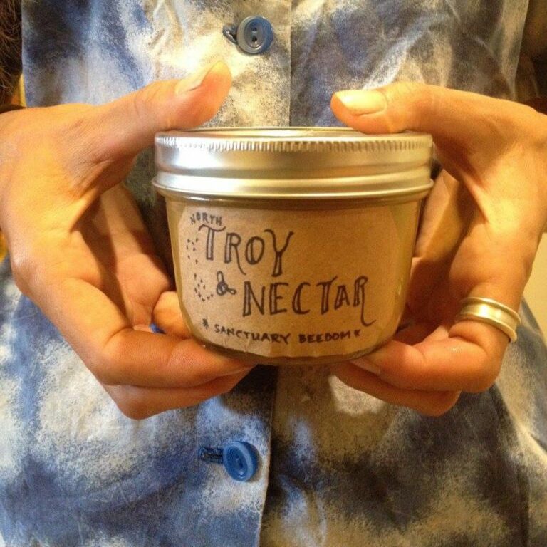 Close up of a small jar of honey being held by two hands. The jar label is hand drawn and reads "Troy Nectar"