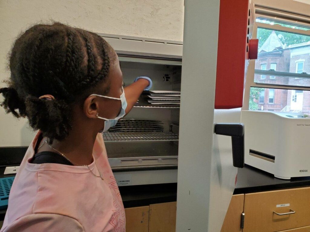 A dark-skinned girl, wearing a surgical mask and gloves, puts water sample packets into the refrigerator at the NATURE Lab. 