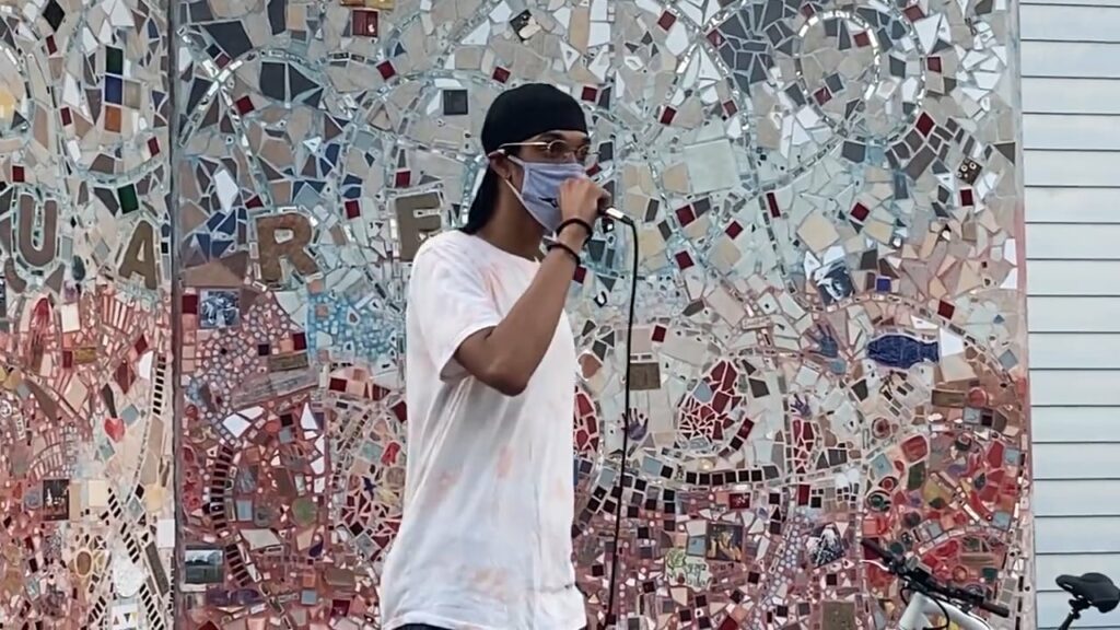 Photo of a person of color wearing glasses standing in front of a mosaic wall, giving a speech.
