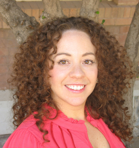 A light-skinned woman with curly brown hair smiles at the camera. 