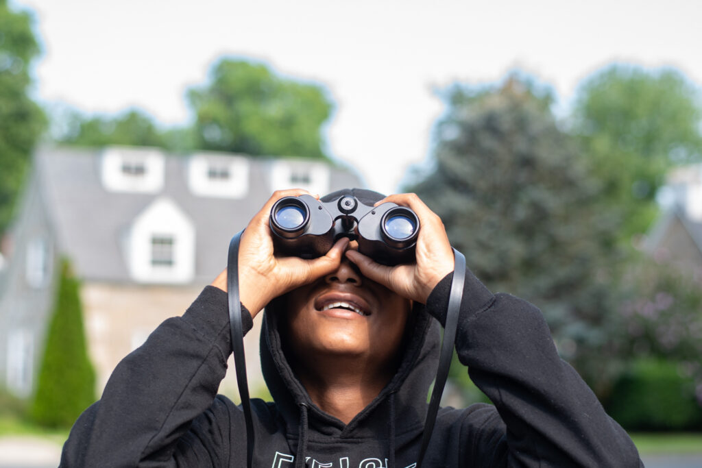 A brown-skinned person wearing a black hoodie smiles and looks through a pair of handheld binoculars. 