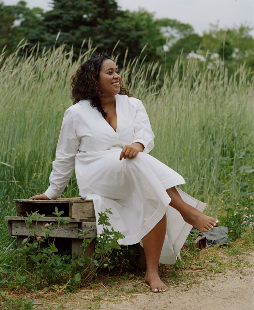 A photoshoot of Mae-ling Lokko wearing a white dress in a flower field, smiling off to the side. She is a person of color with long curly brown hair.