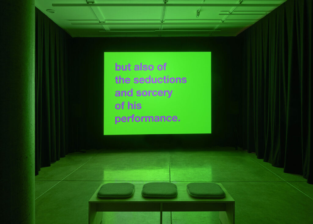 room bathed in green light with a projection of "but also if the seductions and sorcery of his performance."