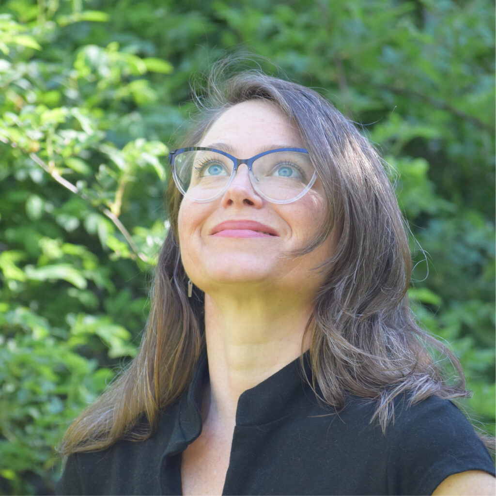 Headshot of Natasha Myers, wearing glasses and looking up while standing in greenery outdoors. 