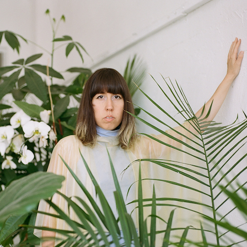 Portrait of Tiare Ribeaux surrounded by tropical plants in an indoor space. Tiare wears a draped and sheet beige top that matches the grown out highlights at the tips of her shoulder-length brown hair with bangs. 