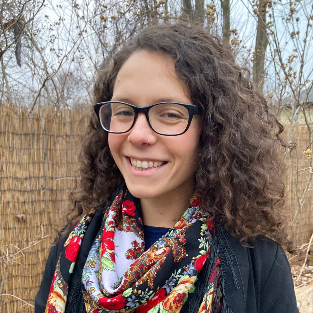 A medium-framed head shot of Ursula Rozum, a light-skinned woman with medium-length curly brown hair, wearing glasses and a floral scarf in front of a fenced forest and smiling for the camera.