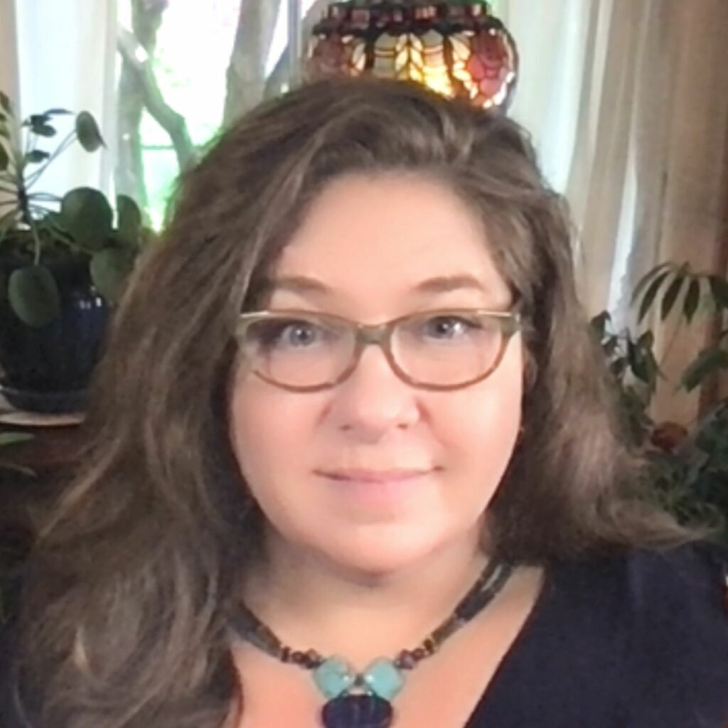 A headshot of Corinne Carey, a light-skinned woman wearing glasses and a turquoise neckless with long, brown hair.