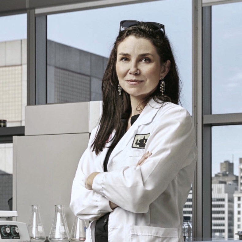 Portrait of WhiteFeather Hunter: wearing a white lab coat while standing with arms crossed in a biology lab with testing beakers.