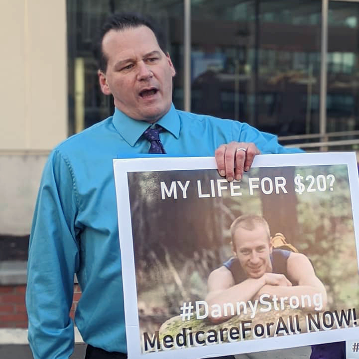 An action shot of Scott Desnoyer, a light-skinned man wearing a light blue button-up and dark blue tie, holding a poster advertising Medicare-For-All with the words My life for $20? #DannyStrong Medicare For All NOW!