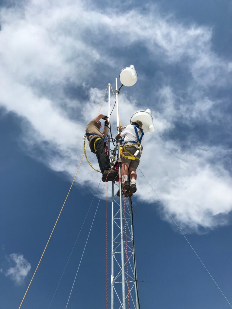 two individuals are standing on a thin radio tower with blue sky and clouds around them