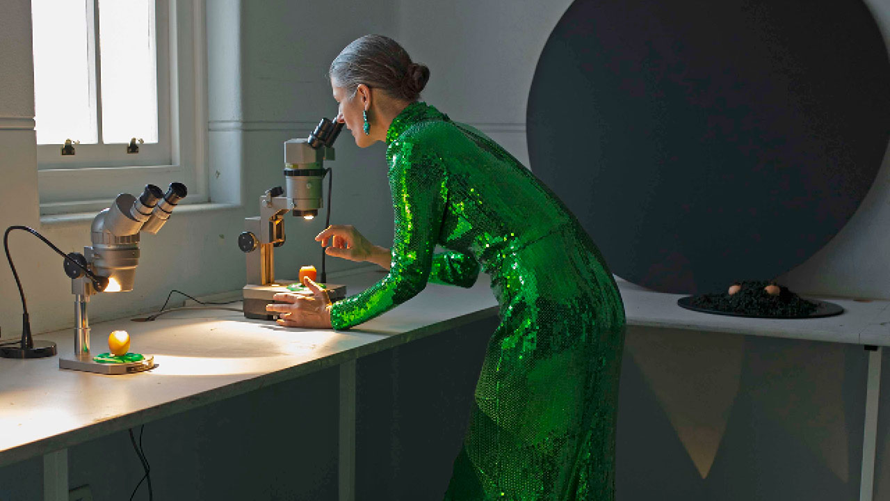 Woman in a green sequenced floor-length dress looking through a microscope in an austere lab.