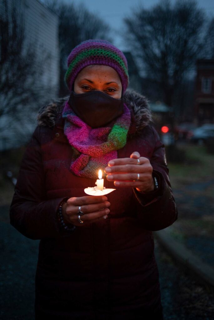 An image of Aileen Javier, a medium-skinned woman, holding a lit candle while bundled up in a multicolor scarf and black jacket, wearing a black face mask while looking at the camera.