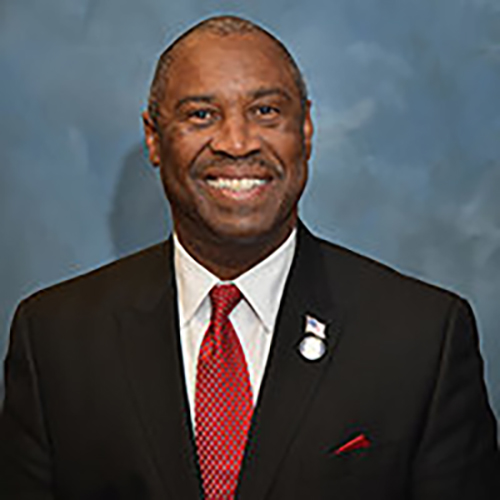 A headshot of Reverend Charles Burkes, who is wearing a black suit with a red tie and smiling for the camera.