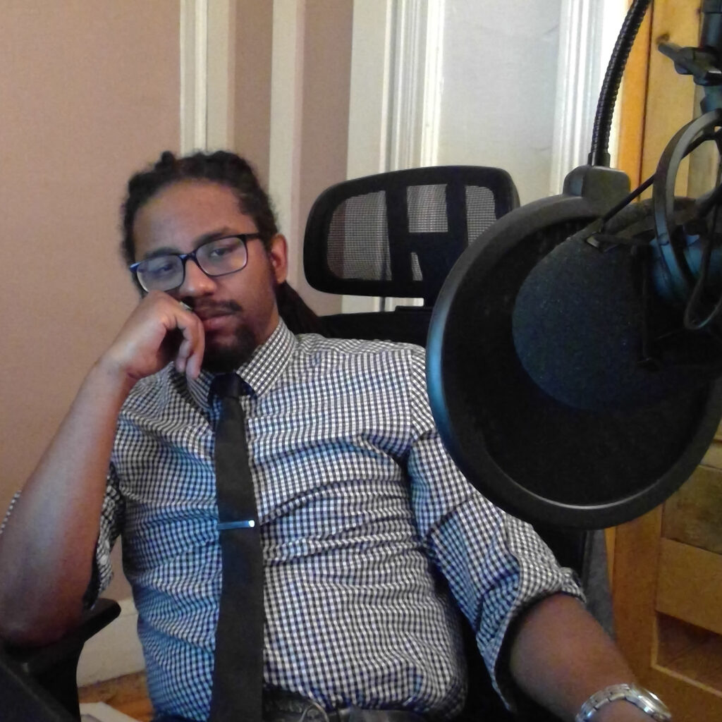 An action shot of Dan Lyles, who seems to be recording a podcast/ participating in an interview. He is wearing a plaid button-up and a black tie.