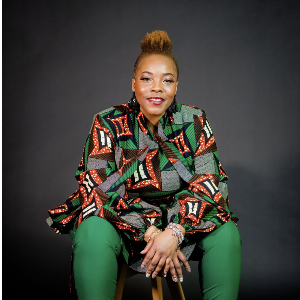 A medium-framed shot of Daquetta Jones, who is wearing an abstract green and red shirt with green pants. sitting on a stool.