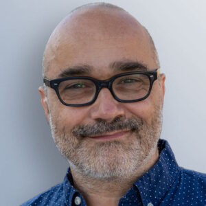A headshot of Dmitri Mugianis, who is smiling for the camera and wearing glasses and a blue button up/
