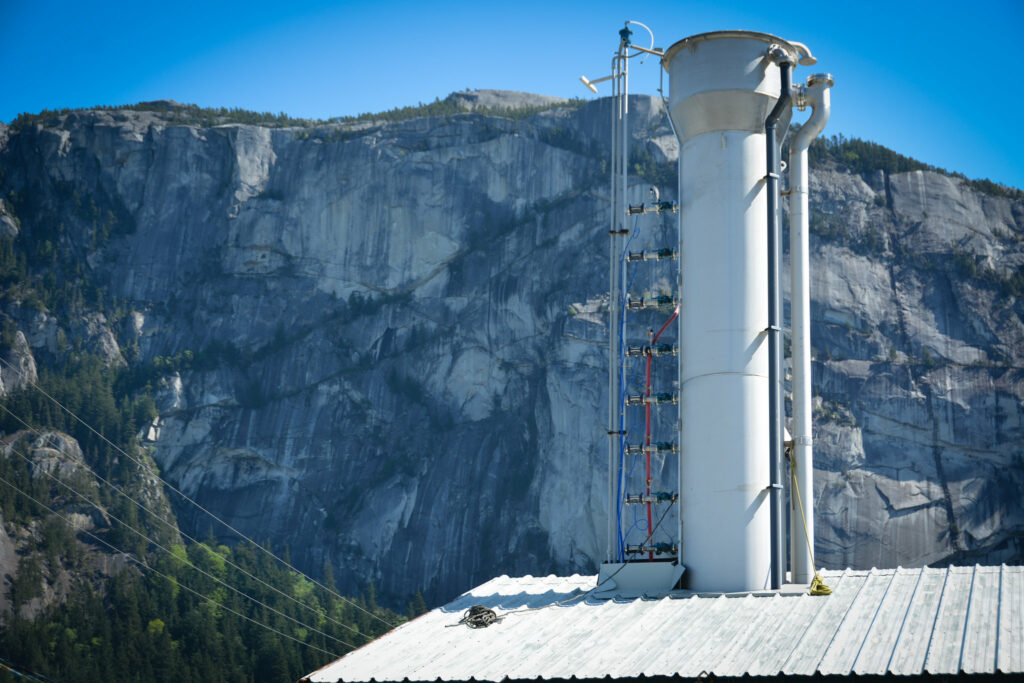 A large, metal stack protrudes vertically from a roof. In the background is a rock face. 