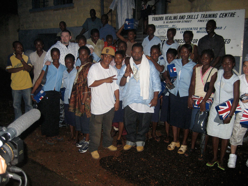 Production still from Bling: A Planet Rock taken by Irena Mihova. A group of African young adults from Sierra Leone pose for a picture with a man in a white shirt making hand signs. A video camera is in the bottom left corner of the photo's frame.