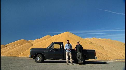 Two men and a black pick-up truck, standing in front of a mountainous amount of corn.