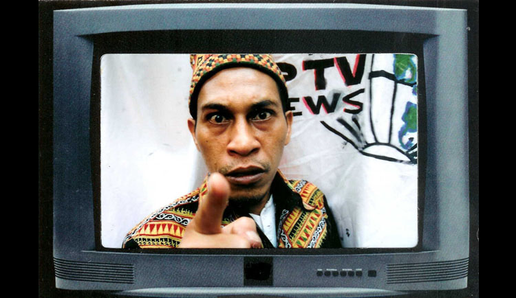 A television monitor with a man in traditional Indonesian shirt and hat is looking into and pointing at the camera. He stands in front of a banner with a globe, with the word news visible.