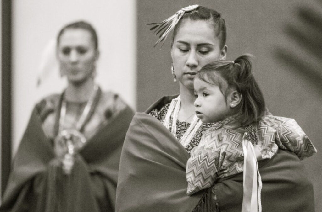 A black and white photograph of two Indigenous women wearing traditional attire. One is holding a small child in her arms.