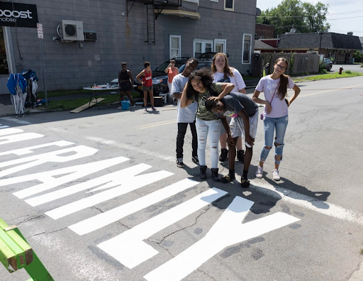 A group of youth from the Sanctuary for Independent Media's 2018 Uptown Summer program. They are gathered in the middle of a street that has a painted message on the road.