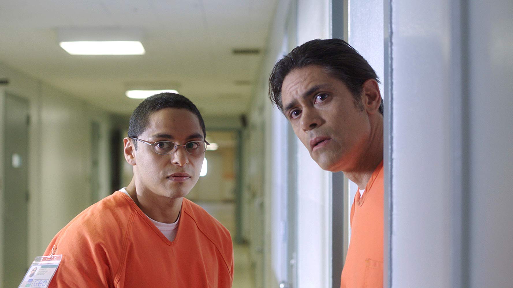 Still from Alex Rivera's film The Infiltrators. Two men in a detention center, wearing orange uniforms, look down a hallway.