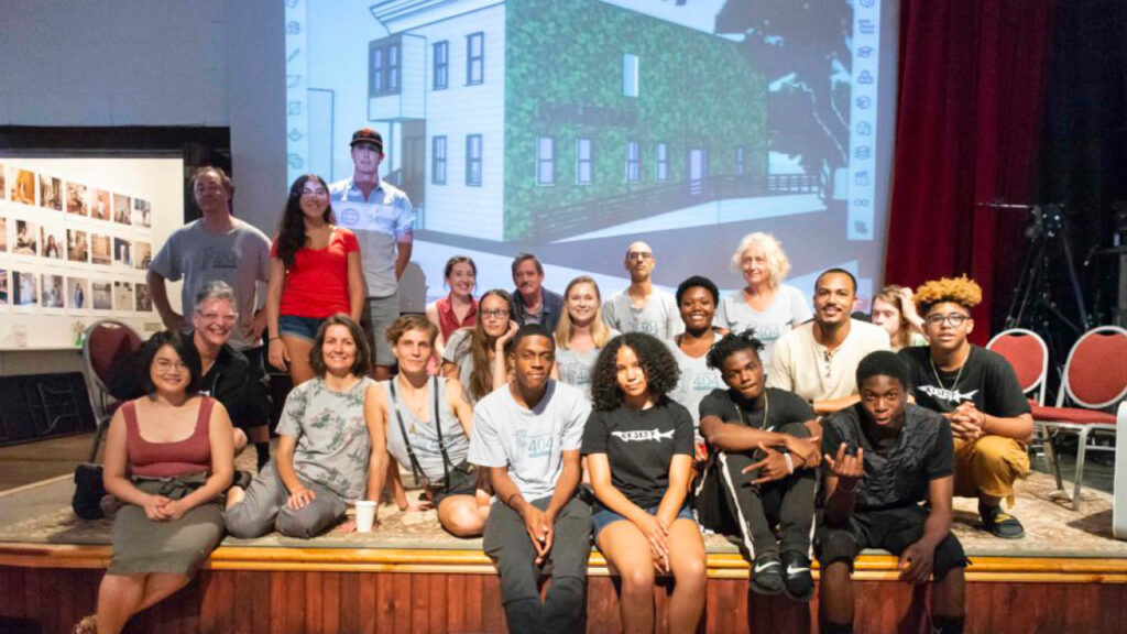 People of various ages and races pose for a group photo on a small stage in front of a black-and-white projection of a two-story urban rowhouse. 