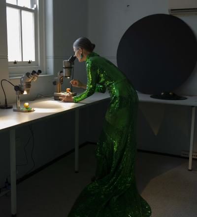 Picture of Kiera O'Reilly, a woman wearing a green floor-length gown, in a laboratory, looking into a microscope. Her hair is pulled back in a bun. The microscope is by a window with sunlight streaming in. 
