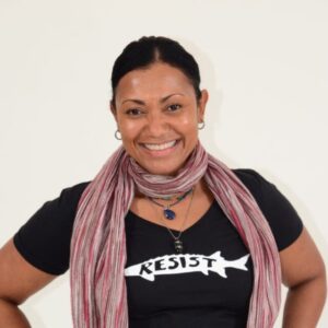 A portrait photograph of Aileen Javier, a latinx woman who is wearing a black t-shirt with a fish that says "Resist" and a pink scarf. Her hair is pulled back, she is wearing earrings, and she is smiling.